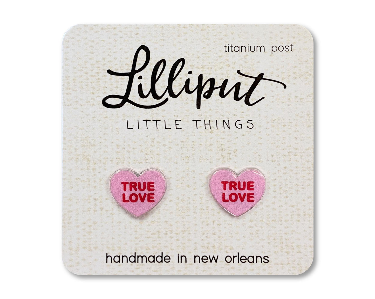 Stud earrings on a card from the brand Lilliput Little Things. The earrings are heart shaped and pink with red text reading "True Love"