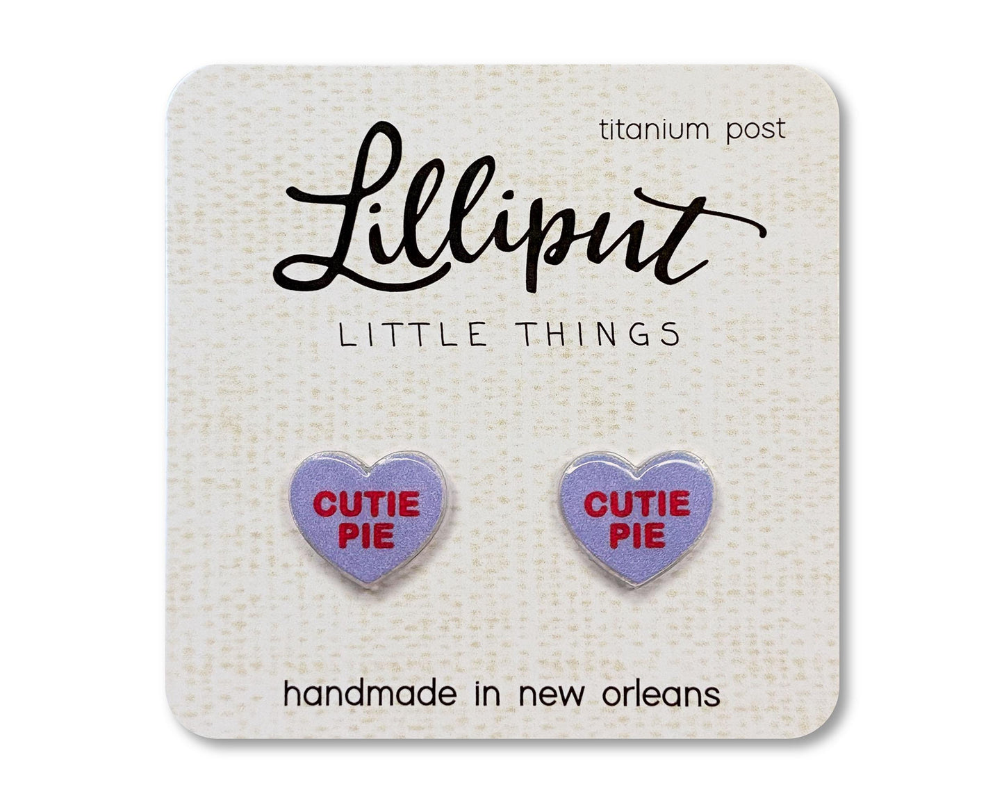 Stud earrings on a card from the brand Lilliput Little Things. The earrings are heart shaped and purple with red text reading "Cutie Pie"