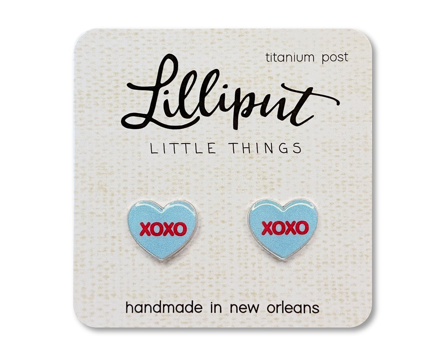 Stud earrings on a card from the brand Lilliput Little Things. The earrings are heart shaped and blue with red text reading "XOXO"