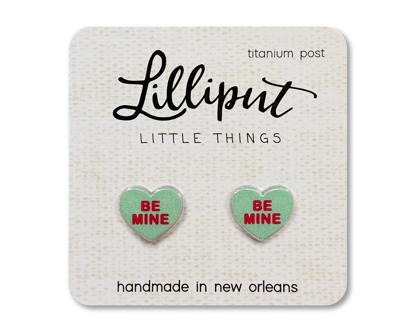 Stud earrings on a card from the brand Lilliput Little Things. The earrings are heart shaped and green with red text reading "Be Mine"