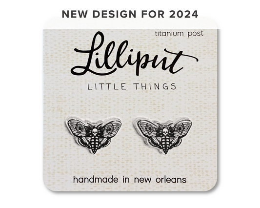 A pair of moth earrings with skull patterns on them, displayed on a card that reads 'Lilliput Little Things, handmade in New Orleans, Titanium Post"